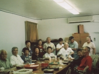 B Company chapter meeting in clubhouse sent to Takeshi Teshima. Sitting around the table from left: Robert Takashige, Thomas Tsubota, Rikio Tsuda, Mrs. Rikio Tsuda, Mrs. Hajime Kodama, Mrs. Sonsei Nakamura, Joichi Muramatsu, Mrs. Raymond Nosaka, Raymond Nosaka.  Standing from left to right:  Mrs. Bernard Akamine, Mrs. Robert Arakaki, Robert Arakaki, Joyce Muramatsu Doi, Bernard Akamine, Drusilla Akamine Tanaka. [Courtesy of Ted Teshima]